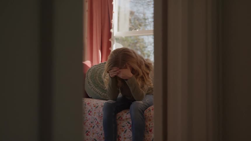 Teenager seated on floral bedspread covers face with hands in room lit by natural daylight