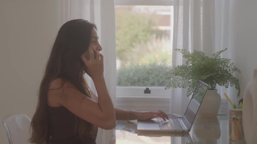 A young woman sits behind a desk in a home office at midday typing on a laptop before picking up her phone to make a call