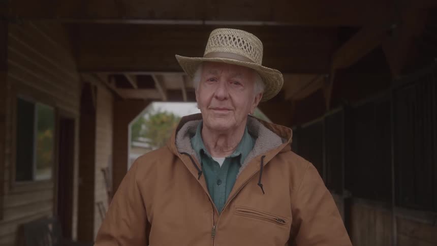 Elderly man wearing a straw hat and brown jacket posing in a rural setting