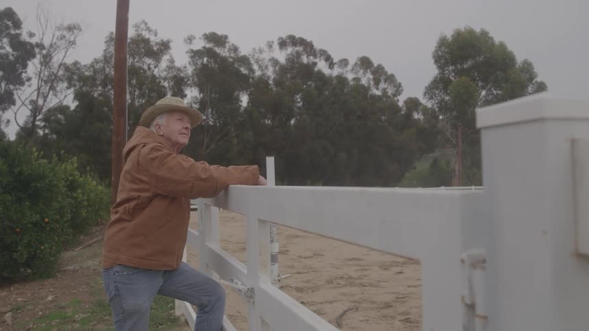 The farmer is leaning against the fence on the ranch