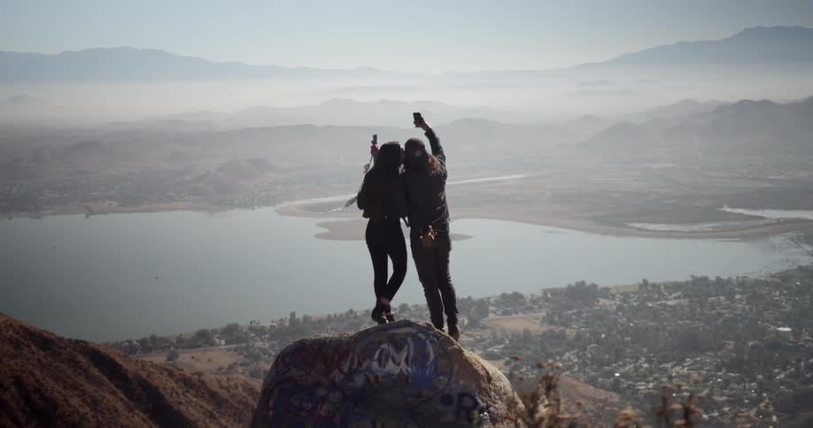 A man and woman standing on a rock high above a lake take a selfie