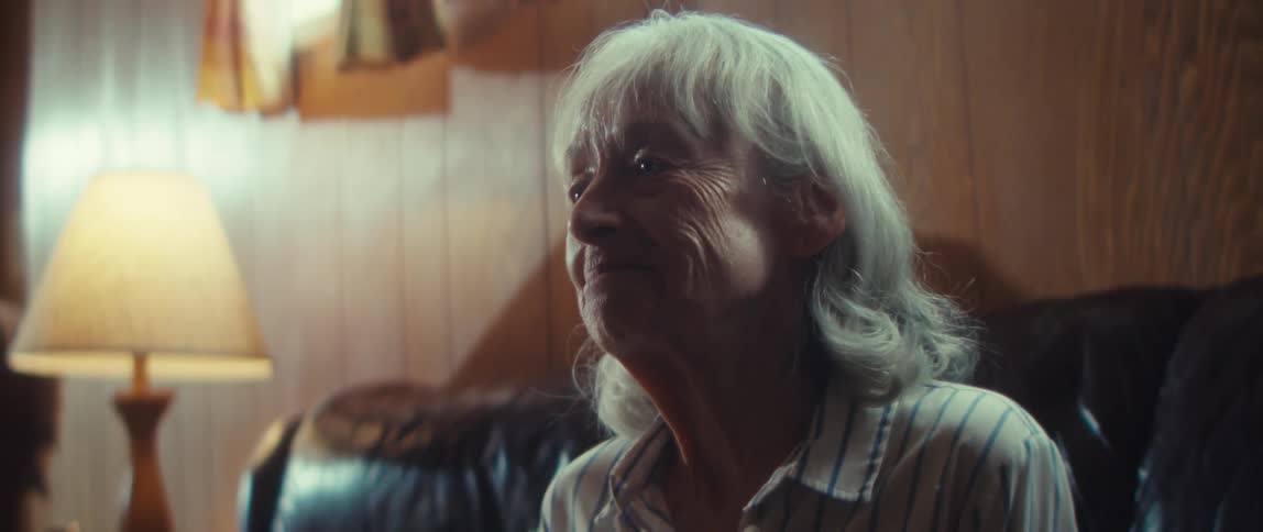 An elderly woman sits on a couch in the living room smiling as she watches tv with a table lamp beside her