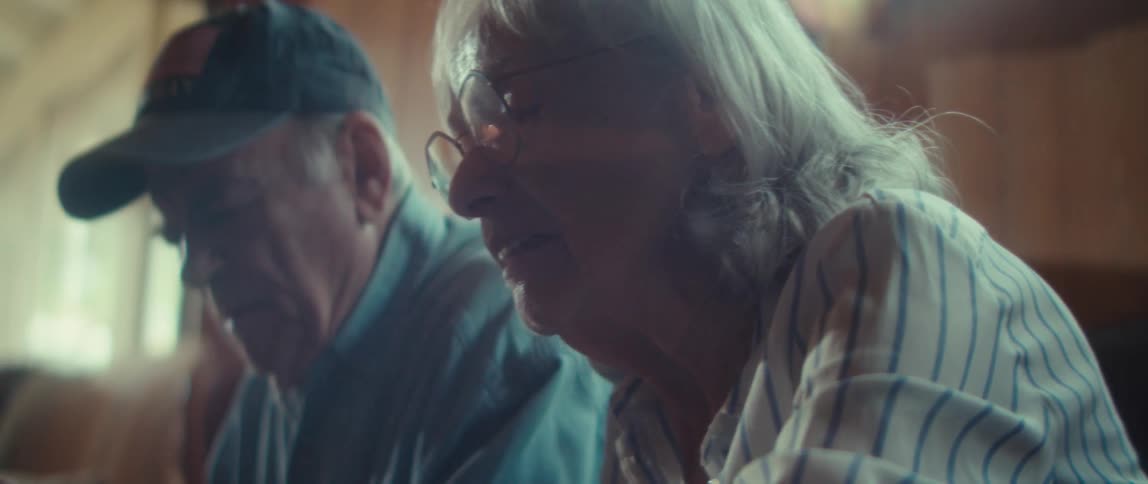 The video is a mediumclose shot of an elderly woman with long gray hair and glasses looking down at something offcamerashe is sitting beside an older man wearing a baseball cap and the room is softly lit with warm light accentuating the wood