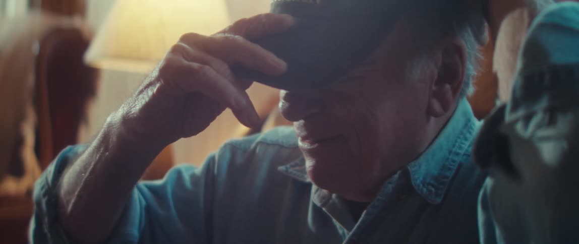 An older man wearing a blue shirt and a cap with an american flag and the word veteran smiles widely while an older woman is visible beside him in soft indoor light