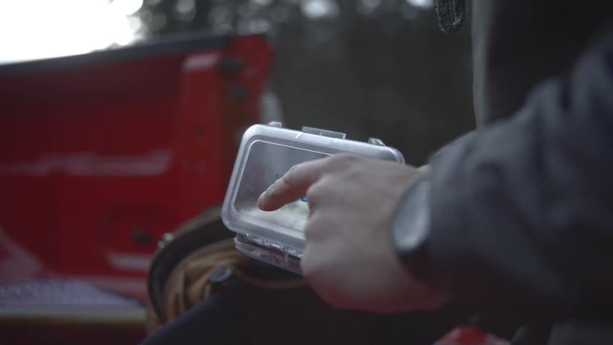 A close view of a mans hands opening a clear plastic box filled with white fishing flies while sitting by the red tailgate of a truck in a dark outdoor setting