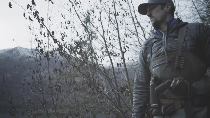 A man wearing a baseball cap and outdoor gear walks alone through a steep sparse and gray winter forest trail before pausing to look out at the cold mountainous landscape