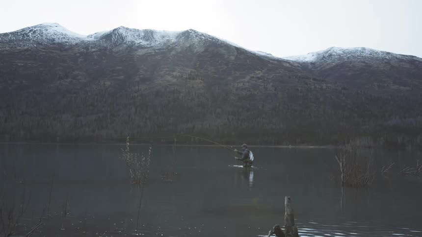 Man standing in a body of water located near a mountainon a winter day casts his line
