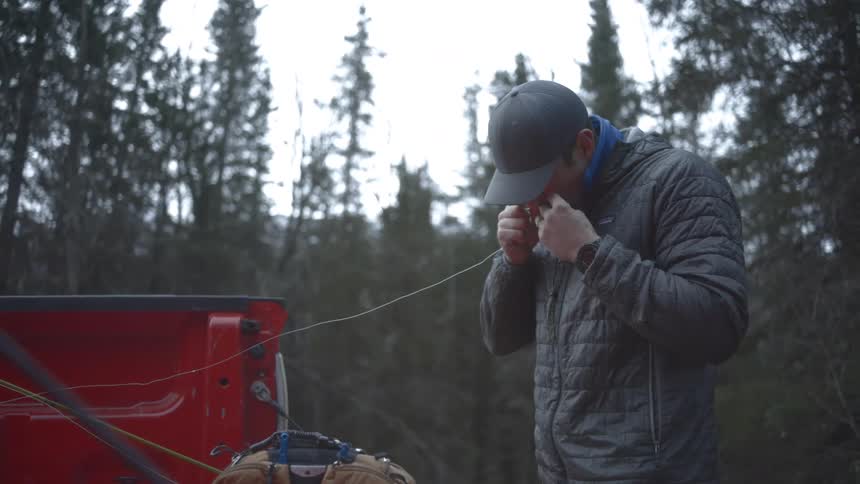 A man in a baseball cap and a puffy gray jacket stands in the misty sparse winter woods next to the red open tailgate of a truck meticulously tying a knot in a thin line
