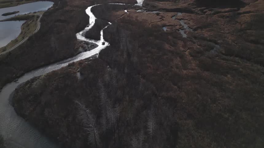 An aerial view of a winding river with silvery water slowly descends revealing a vast dark brown mountain valley with patches of marshy water and the bright sun peaking over a high peak