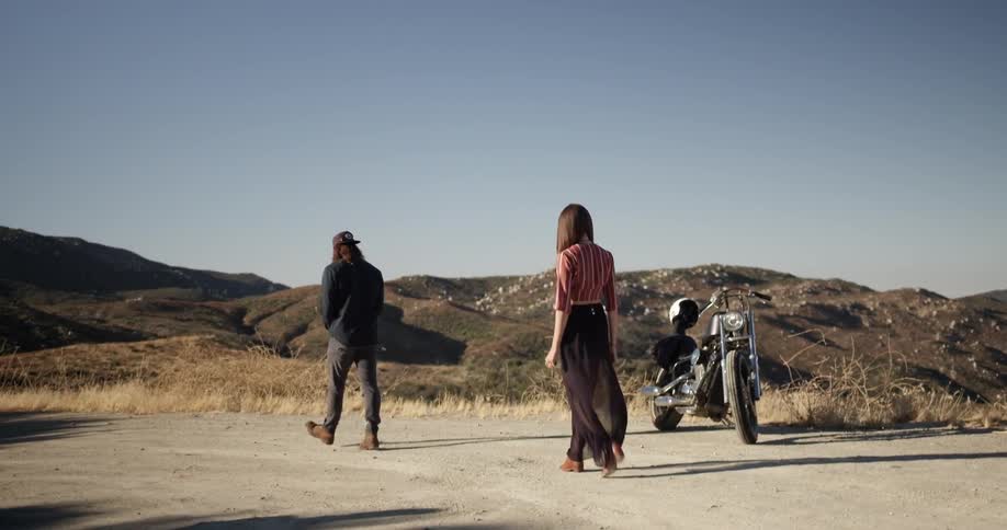 A man and woman walk by a parked motorcycle in a rural area