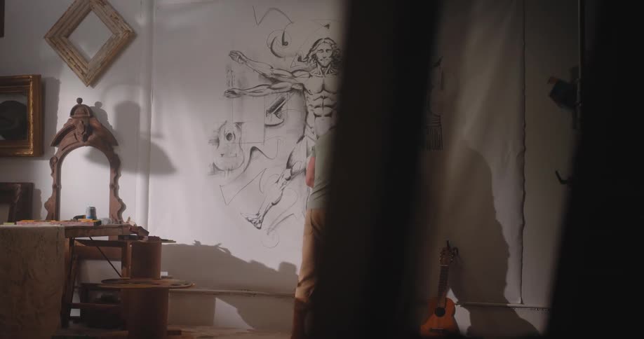 A muscular man in a green shirt and tan pants stands in a rustic art studio drawing a large anatomical male figure on a white wall framed by a dark foreground and antique furniture