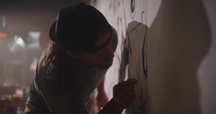 Close up of a young male artist finishing a large drawing on a white canvas on the wall in a room during daytime
