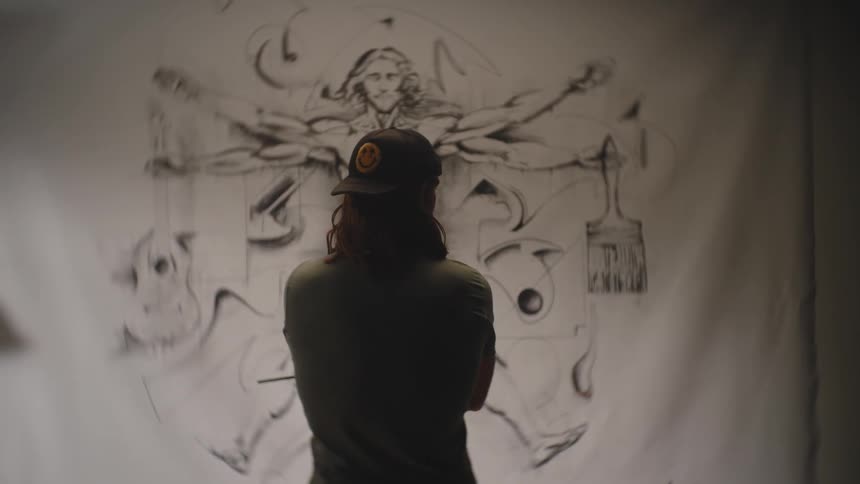 A man with long hair and wearing a backwards cap stands with his arms crossed looking at a hanging sheet with a painting based on the vitruvian man but with objects hanging from the hands