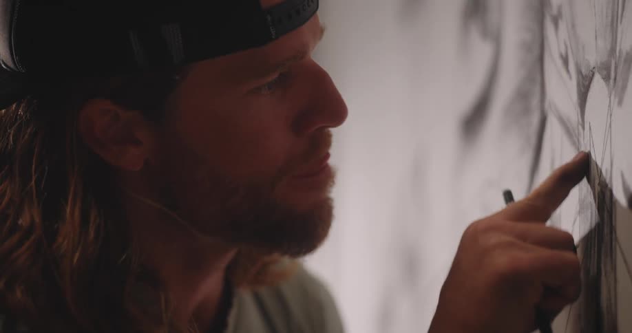A close profile view shows a redbearded man wearing a backward baseball cap pointing his finger at the large black drawing on a bright white wall he is creating