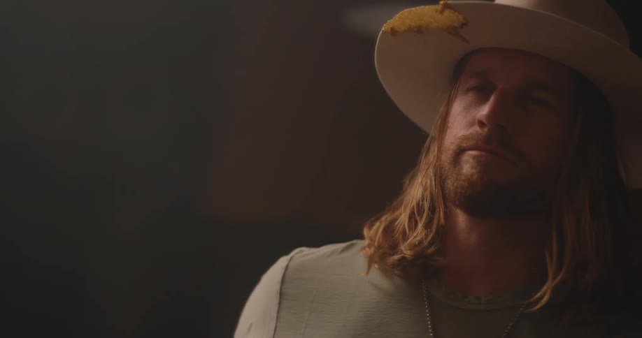 A man with long hair and a beard adjusts his hat and looks around with a contemplative expression in a dimly lit setting