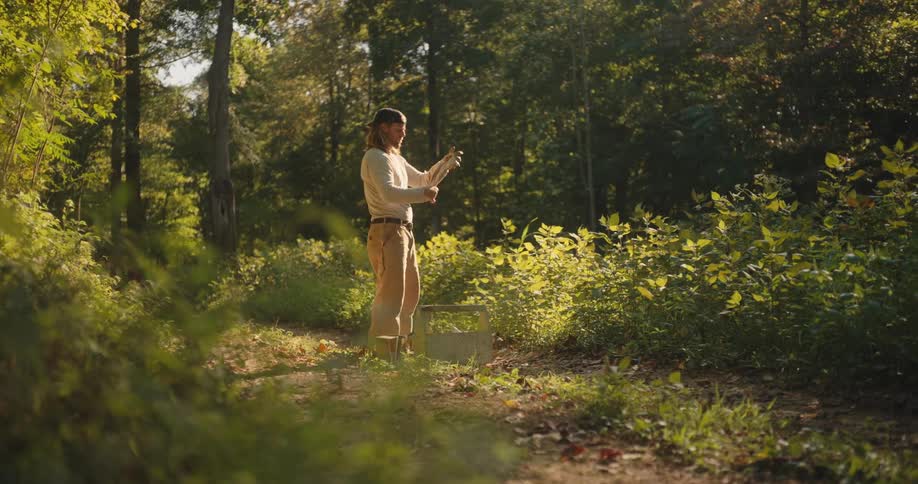 A man with long hair wearing a white long sleeved shirt tan pants and a baseball cap is standing on a dirt path in a sunlit forest putting on work gloves from his hands