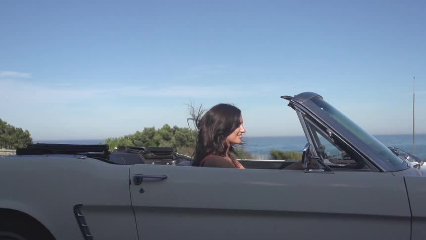 A young, brunette woman driving a white convertible next to the ocean