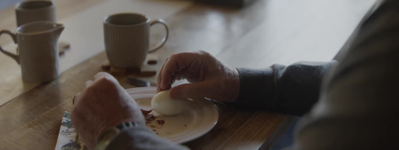 Mans hands peeling a boiled egg over a plate on a breakfast table