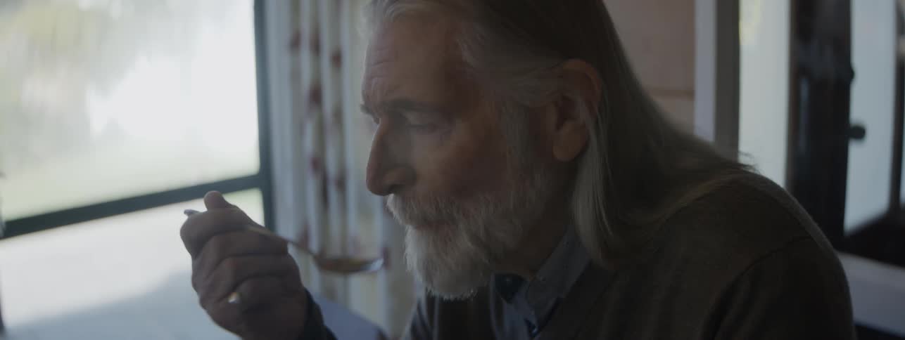 An older man with long gray hair and a long gray beard who lifts a spoon to his mouth to eat food