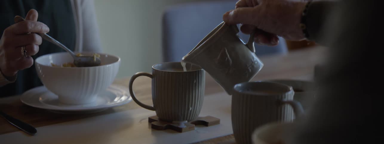 A hand holding a vessel and pouring a liquid into a ribbed mug sitting on a wooden coaster on a table