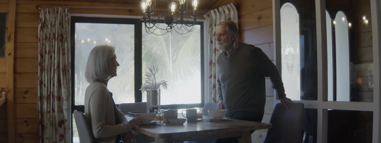 An older man adjusts the chair of a woman of the same age sitting at a breakfast table and walks around the table to sit himself while they both smile and talk then the woman reaches out and holds his hand across the table