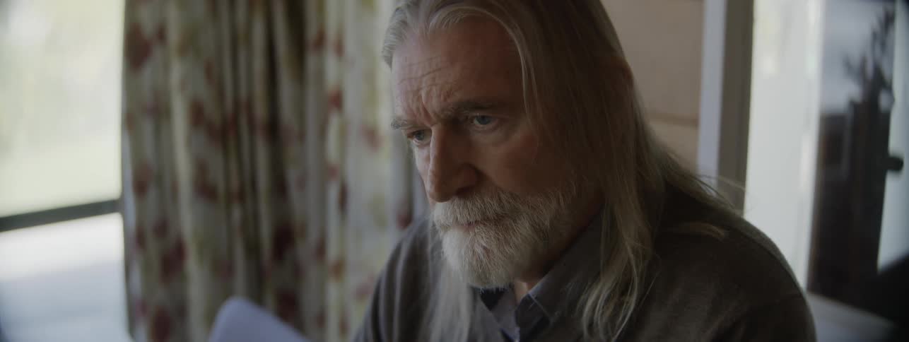 Head and shoulders of an older man with a long gray beard and long gray hair who is sitting and looking down at a surface