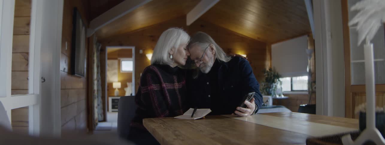 A senior man and woman with white hair sit close together at a wooden table in a cozy cabin interior looking down at a smartphone and a small notebook before touching their foreheads together