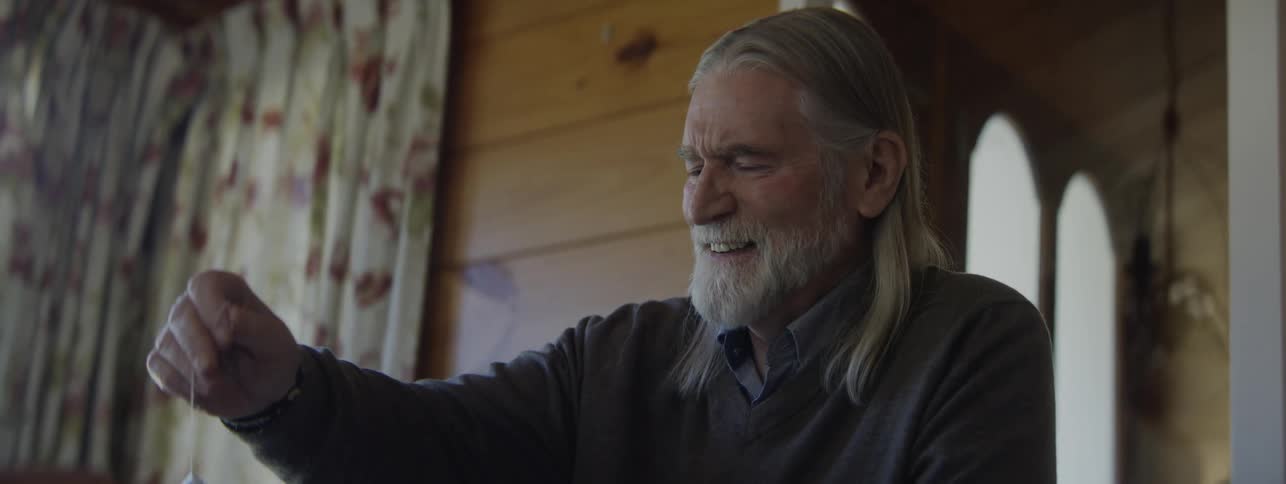 A long haired bearded older man smiles and talks while gesturing appearing both happy and a bit distressed