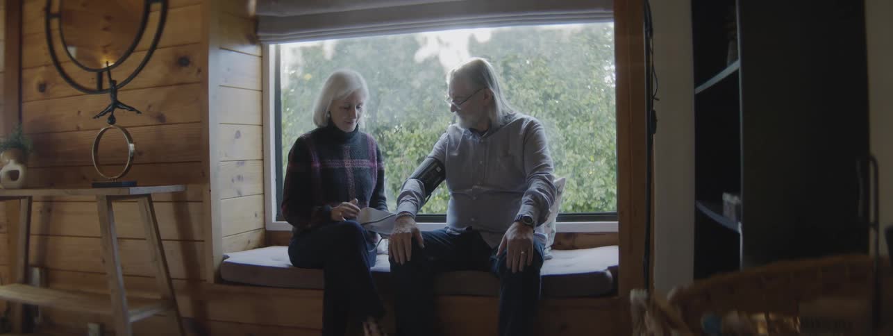The video is a midshot of an older couple sitting on a window seat in a woodpaneled roomthe woman is wrapping a blood pressure cuff around the mans arm suggesting she is taking or preparing to take his blood pressure