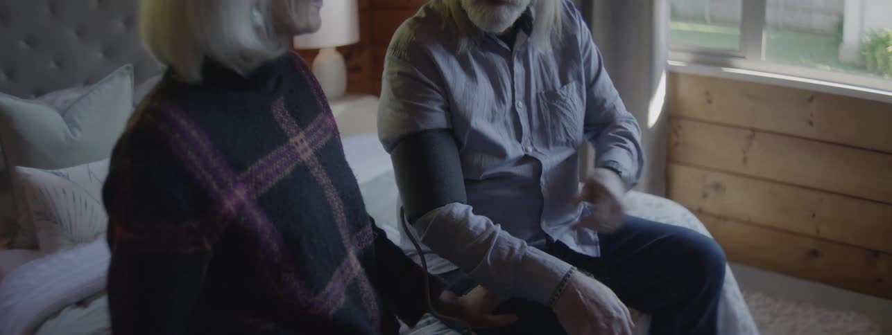 A senior man with long white hair and a gray shirt sits on a bed with a senior woman in a dark patterned top who operates a blood pressure cuff on his arm in a bright modern room