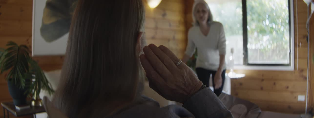 The video is an overtheshoulder shot of one woman speaking to another in a brightly lit room with woodpaneled wallsthe woman in the foreground has her hand raised to the speakers face in a gesture of gentle comfort or concern