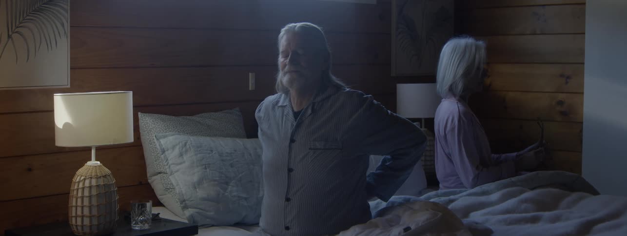An older man with a long white beard sits up in a darkened woodpaneled bedroom clutching his lower back in pain while a woman sits beside him looking away