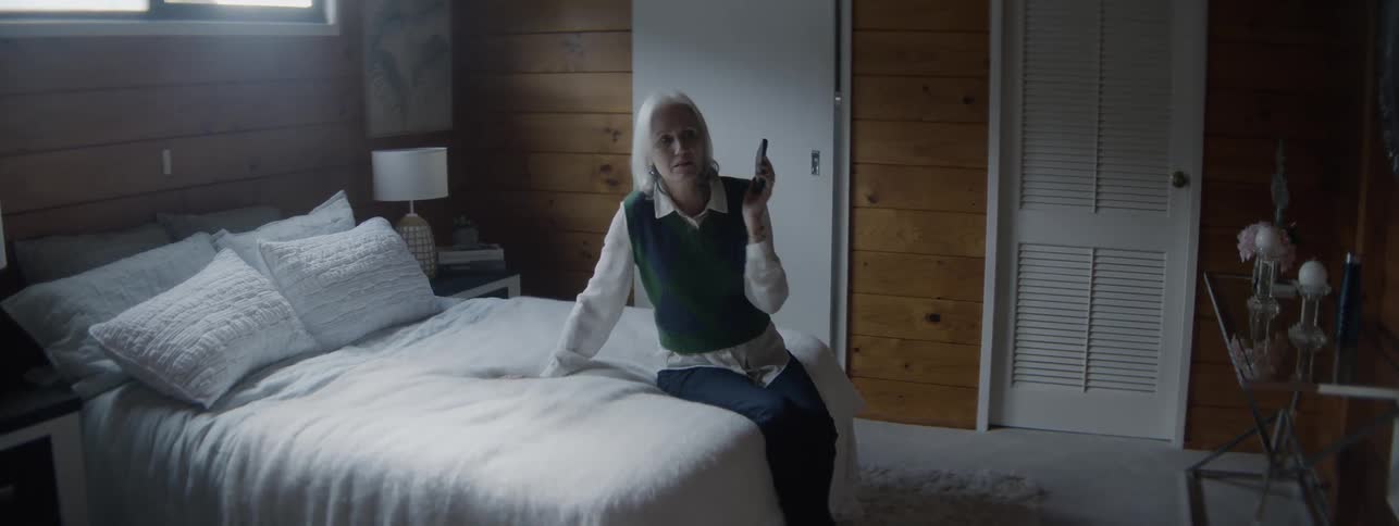 A middle aged woman making a phone call while sitting on a bed in a bedroom during daytime rushes to check who is at the door