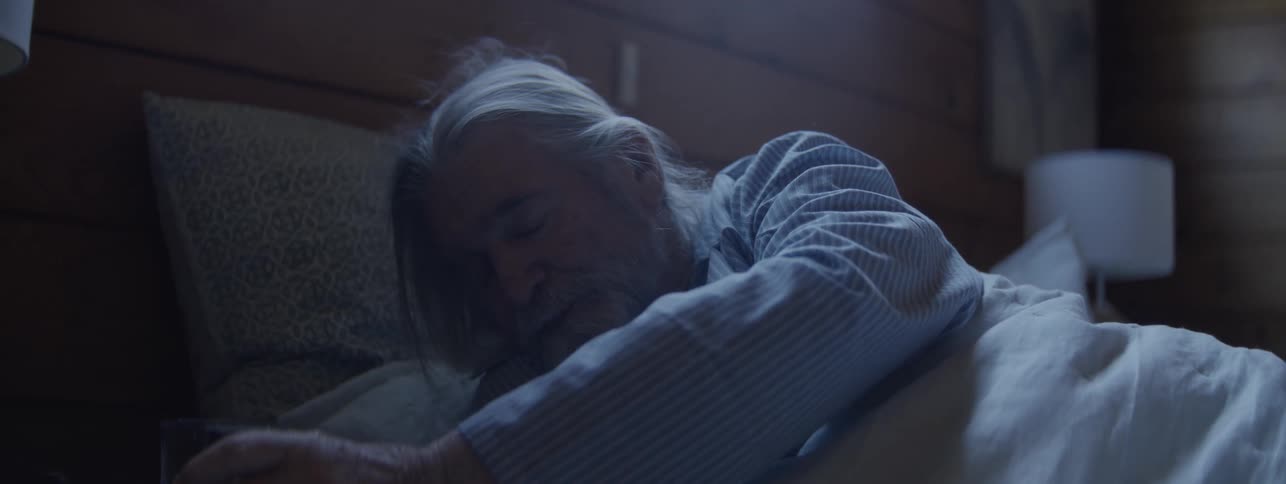 An older man with shoulder length hair tossing and turning in bed gets up just enough to take a sip of water from a glass on his nightstand then goes back down to bed