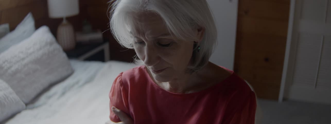 An elderly woman with grey hair sits on the edge of her bed at midday wearing a red dress and filled with sadness