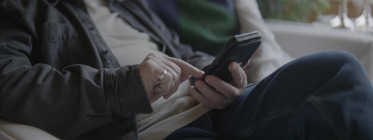 Mans hands using a smartphone in an indoor setting while the man is sitting on a couch with a person on his side
