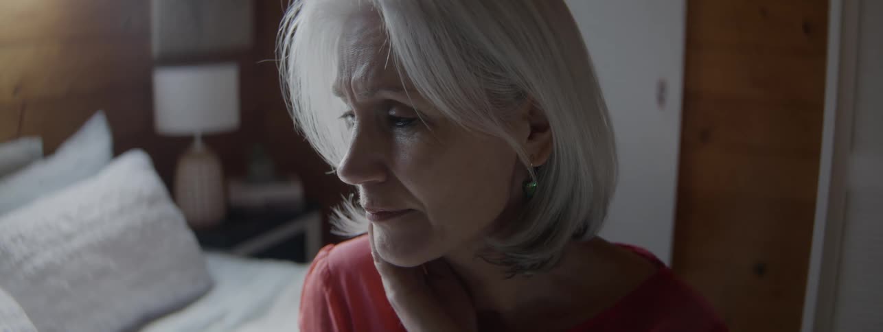 An older woman sits in a bed in a room with one hand in her chin