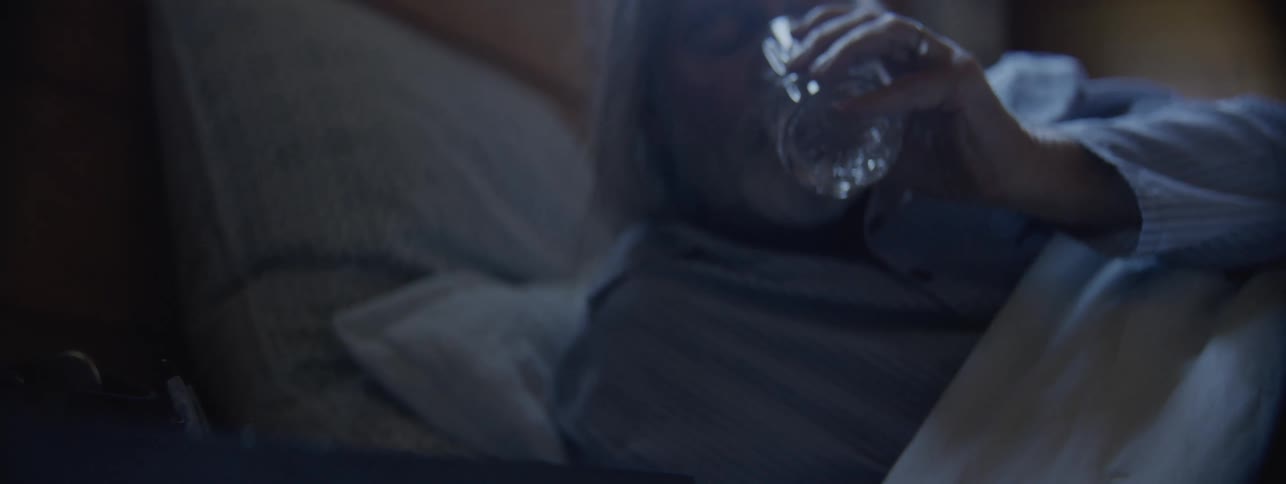 The video is a lowlight closeup shot of an older man waking up in bedhe reaches for and drinks from a cutcrystal glass of water sitting on his bedside table before lying back down