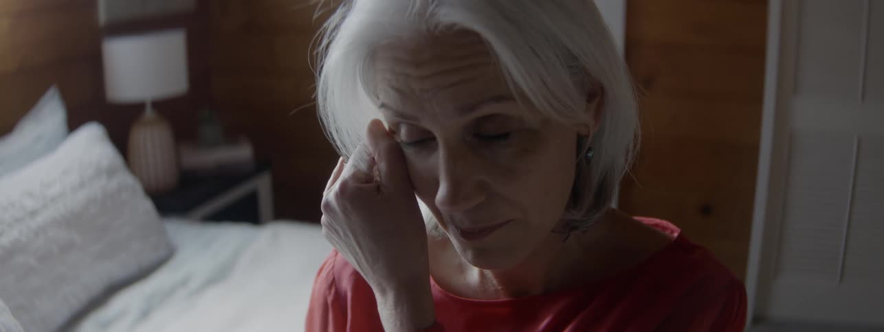 An older woman with shoulder length hair sitting at the edge of a bed looks sad as she uses her thumb to wipe a tear from the corner of her eye