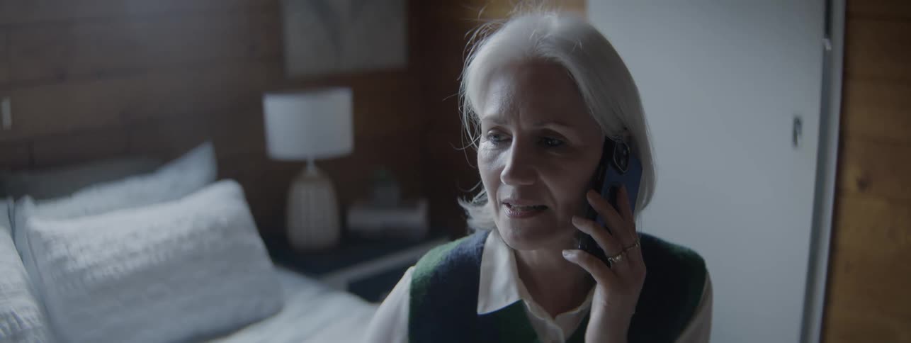 Elderly woman holds a blue phone and talks on a call inside a bedroom