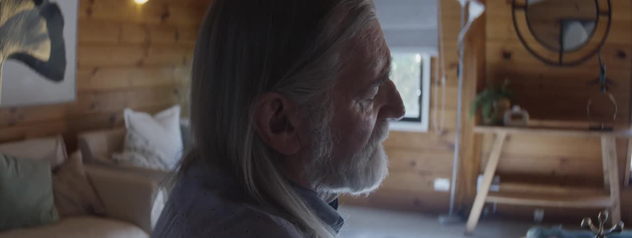 An older man with long hair sitting in a living room frowns in pain