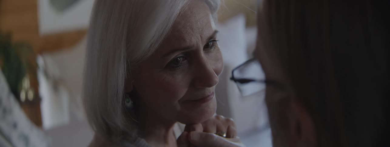 The face of an older woman with short gray hair looking at a second person whose face is out of focus in the foreground as the two people hold hands and the man leans in