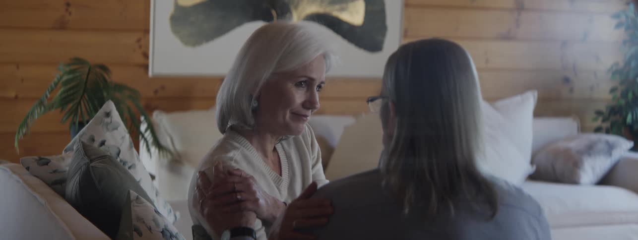 The video features two elderly women having an intimate conversation in a cozy living space with wood panelingthe woman with short silver hair on the left is holding and gently comforting the woman on the right by grasping her arms and shoulder
