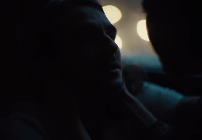 A distressed male grimaces in dim light as another person grabs him with blurred lights glowing behind
