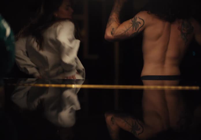 A longhaired man with tattoos is shirtless and dancing beside a woman in a white sweater who is leaning on a reflective black table in a darkened room before they move closer to kiss