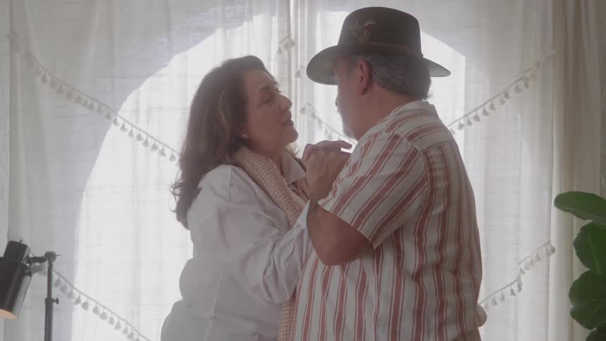 A man wearing a hat and a striped shirt slow dances while embracing a woman in a light shirt standing close and looking into each others eyes