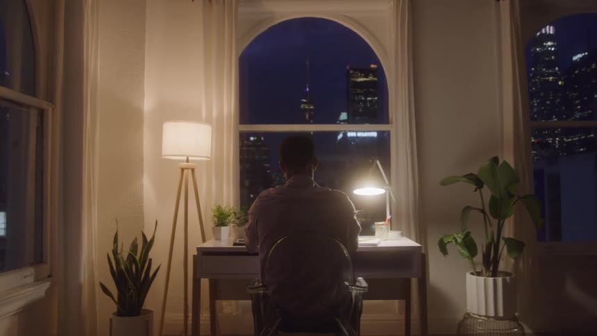 A man sitting at a desk by a window at night stretches with his hands behind his head than relaxes into a straight position