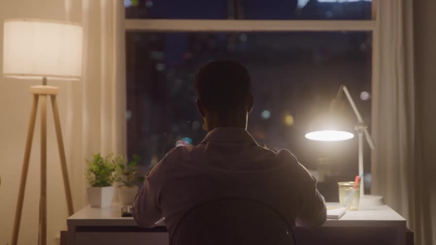 The back view of a man seated at a desk in a dimly lit room with a window overlooking a dark city stretching his arms behind his head and then leaning forward to work at night