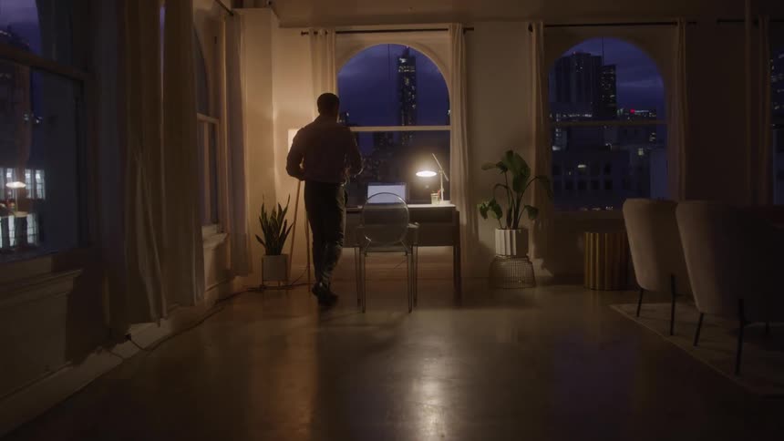Man about to sit in front of a laptop in a softly lit room at night with an urban view