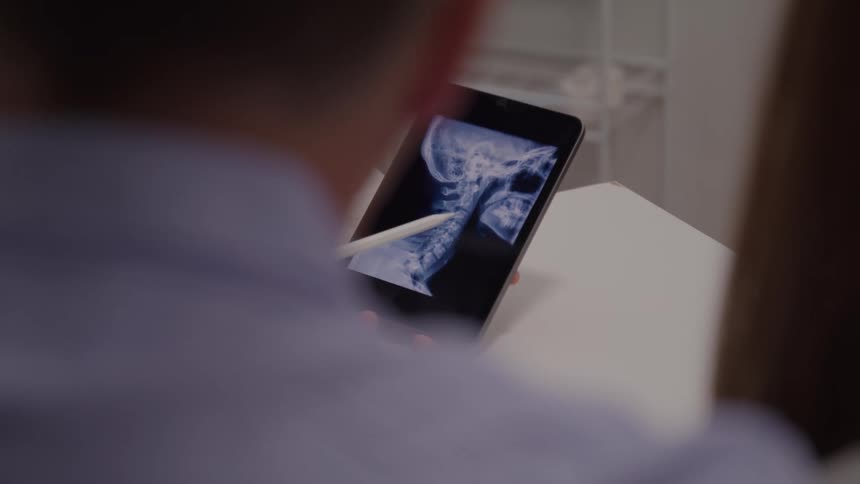 A doctor holds a tablet in his office showing a patient an xray image of a human skull displayed on the screen